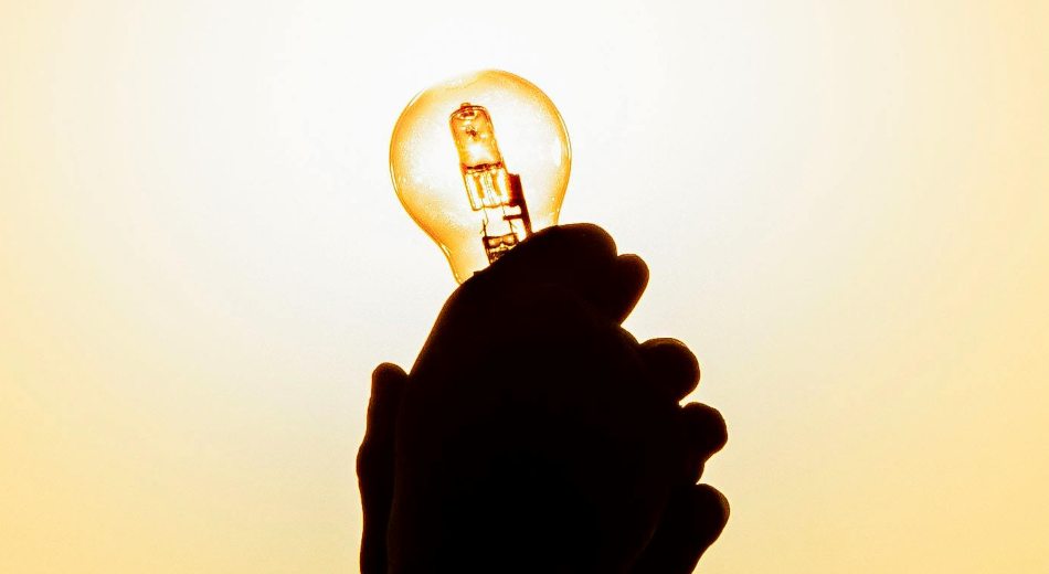 Esta es una descripción corta de la imagen:  Esta es una imagen a contraluz (silueta) dominada por tonos cálidos, amarillos y dorados. Muestra la silueta de una mano que sostiene una bombilla encendida, la cual irradia luz intensamente. Es un símbolo clásico de la inspiración o una nueva idea.